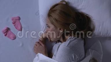 哭泣的女士躺在床上，手里拿着粉红色的婴儿袜子，产后抑郁